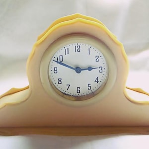 May include: A vintage-style mantel clock with a cream-colored face and a light yellow frame. The clock face has black numerals and blue hands. The clock is designed with a decorative, scalloped top and a rectangular base.
