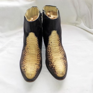 以下が含まれることがあります： 前面と側面にヘビ柄模様のある黒いアンクルブーツのペア。ブーツはポインテッドトゥで、側面にジッパーが付いています。内側はタン色の素材で裏打ちされています。