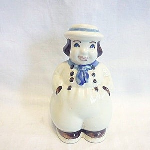 Puede incluir: Una figura de cerámica de un marinero con un atuendo blanco, una bufanda y un sombrero azules. La figura tiene cabello y zapatos marrones, y botones negros en la parte delantera. El marinero tiene una cara sonriente y está de pie.