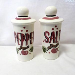 May include: A pair of vintage ceramic salt and pepper shakers. The white shakers have a rounded top and are labelled "Pepper" and "Salt" in burgundy lettering. Each shaker is decorated with a cherry and leaf design.