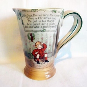 Könnte beinhalten: Vintage Keramikkrug mit cremefarbenem Körper und breitem Henkel. Der Krug zeigt eine gemalte Illustration eines Kindes und das Gedicht "Little Jack Horner". Der Griff ist mit grünen Zierleisten versehen, und der Sockel ist hellgelb.