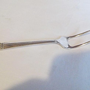 May include: A silver-toned metal serving fork with two long, slender tines. The handle features a rectangular end with an intricate, embossed floral pattern, transitioning into a smooth, tapered neck leading to the fork head.