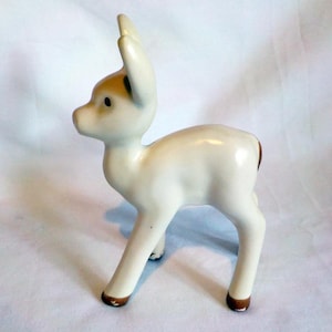 May include: A small ceramic figurine of a standing fawn with a smooth, off-white glazed finish. The deer features pointed ears, a dark brown nose, dark eyes, and small brown accents on the tips of its hooves. It stands in a profile view.