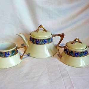 Puede incluir: Un juego de té color crema con detalles dorados. El juego incluye una tetera, una jarra para la crema y un azucarero. Cada pieza está decorada con un patrón floral azul y verde alrededor del borde. La tetera tiene un pico curvo y un asa dorada.