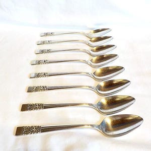 May include: Eight silver spoons displayed diagonally. Each spoon features a rounded bowl and a long, slender handle with a floral design at the end. The spoons are presented on a white surface.