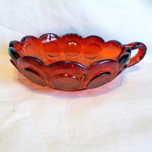 Könnte beinhalten: Eine bernsteinfarbene Glasschale mit Griff. Die Schale hat einen gewellten Rand und kreisförmige Muster am Boden. Der gebogene Griff ist an der Seite befestigt. Diese Vintage-Schale ist wahrscheinlich zum Servieren oder Ausstellen gedacht.