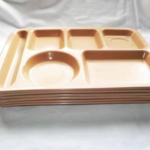 May include: A stack of beige plastic cafeteria trays. Each tray has multiple compartments of various shapes and sizes, including a circular area and several rectangular sections. The trays are stacked neatly, showing their edges.