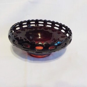 May include: A dark red glass bowl with a decorative, openwork rim. The bowl has a rounded shape and sits on a small, circular base. The rim features a series of evenly spaced, rectangular cutouts. The bowl is likely vintage or antique.