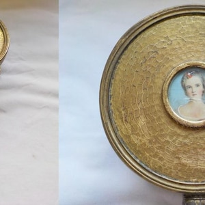 Op de afbeelding: Antieke handspiegel met een ronde, gehamerde goudkleurige lijst. De spiegel heeft een klein portret van een vrouw in het midden. De handgreep is zilverkleurig met sierlijke details. Een vintage item.