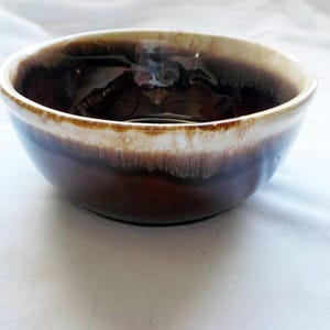 Puede incluir: Un cuenco de cerámica con un esmalte marrón y beige. El cuenco tiene una forma redondeada con un borde ancho. El interior es de color marrón oscuro y el borde tiene un color beige más claro. El cuenco está sobre una superficie blanca.