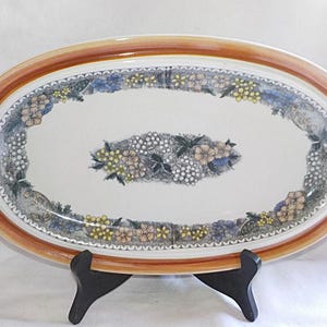 May include: An oval ceramic serving platter with a brown rim and a floral design. The design features white, yellow, and blue flowers and leaves.