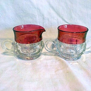 以下が含まれることがあります： ガラス製のクリーマーと砂糖入れの2点セット。縁は濃い赤色で黒いトリムが付いています。透明なガラスの本体には装飾的な模様と小さなハンドルが付いています。