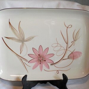 May include: Rectangular serving platter with a cream-colored background and a brown rim. The platter features a floral design with a pink flower and brown and gray leaves and stems. A decorative piece for serving food.
