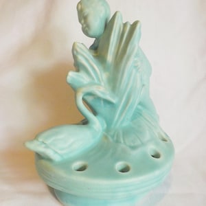 May include: A light blue ceramic flower frog featuring a sculpted figure holding a large fan, accompanied by a swan resting on the base. The circular base includes several evenly spaced holes for arranging stems, with a smooth, matte finish.