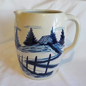 May include: A cream-colored ceramic pitcher featuring a hand-painted blue landscape design. The illustration depicts a small house, evergreen trees, and a winding fence path. The pitcher has a curved handle and a wide pouring spout at the top.