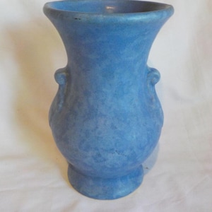 May include: A medium blue ceramic vase with a mottled, textured finish. The vessel features a wide, flared rim, a rounded body, and two small, decorative scroll-shaped handles on opposite sides, resting on a circular pedestal base.