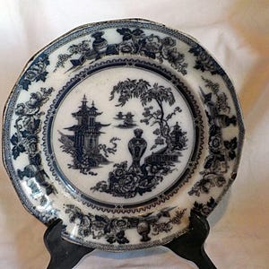 May include: An antique white and blue decorative plate with an Asian-inspired landscape design. The plate features a central scene with a pagoda, trees, and floral border. The plate is displayed on a black stand.