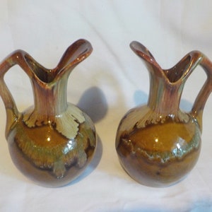 Könnte beinhalten: Zwei Keramikvasen mit bauchigem Boden und geweitertem, offenem Oberteil. Die Vasen haben einen Henkel und sind in Braun-, Grün- und Beigetönen glasiert. Das Design umfasst blattartige Akzente in der Nähe der Oberseite. Die Vasen stehen auf einer weißen Oberfläche.