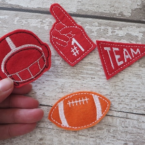 May include: Four felt patches with an American football theme. The patches are red and orange with white stitching. The patches include a football helmet, a football, a foam finger with the number one, and a pennant with the word "TEAM".