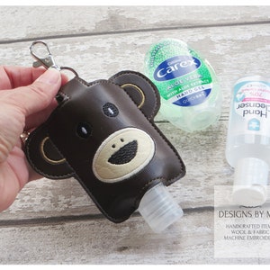 Puede incluir: Porta-gel hidroalcohólico de cuero marrón con un diseño de cara de oso. El porta-gel tiene un llavero plateado y está unido a una botella de plástico transparente de gel hidroalcohólico.