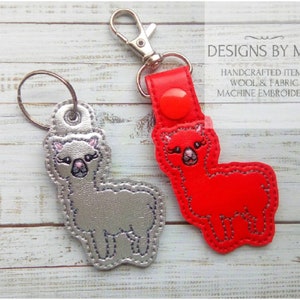 May include: Two alpaca keychains, one silver and one red, with a key ring attached. The keychains are handmade and feature machine embroidery. The text "DESIGNS BY MLP" and "HANDCRAFTED ITEMS WOOL & FABRIC MACHINE EMBROIDERY" is visible in the image.