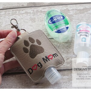 May include: A gray fabric hand sanitizer holder with a brown paw print and the text "Dog Mom" in black with a red heart. The holder has a silver key ring and a clear plastic hand sanitizer bottle inside.