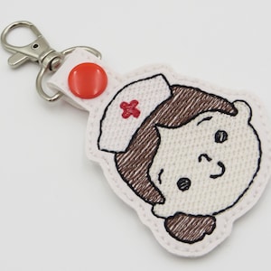 May include: A nurse-themed keychain featuring an embroidered design of a smiling nurse's face with brown hair and a white cap with a red cross. The keychain has a silver clasp and a red button.