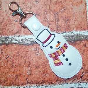 May include: White leather keychain with an embroidered snowman design. The snowman has a red and orange scarf, black eyes, and a red and white hat.