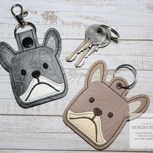 May include: Two gray and brown leather keychains with embroidered French Bulldog faces. The keychains have a metal ring and a snap closure. The text "DESIGNS BY MLP" is visible in the bottom right corner of the image.
