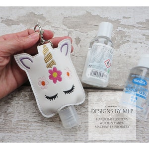 Può includere: Porta-gel disinfettante per le mani a forma di unicorno bianco con un corno dorato e fiori rosa. Il porta-gel è attaccato a un portachiavi argentato. Il porta-gel è riempito con una bottiglia di gel disinfettante per le mani trasparente. L'immagine mostra anche due bottiglie aggiuntive di gel disinfettante per le mani.