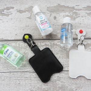 May include: Two black and white leather keychains with a loop and a pocket for a small bottle of hand sanitizer. The keychains are on a light wood surface with three small bottles of hand sanitizer.