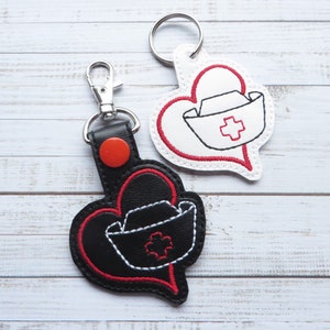 Puede incluir: Dos llaveros en forma de corazón con un diseño de gorro de enfermera. Un llavero es blanco con costuras rojas y el otro es negro con costuras rojas. Ambos llaveros tienen una cruz roja en el gorro de enfermera.