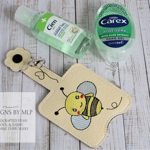 May include: A beige fabric hand sanitiser holder with a bee design and a flower-shaped keyring. The holder is designed to fit a small bottle of hand sanitiser. The text "DESIGNS BY MLP" is visible in the image.