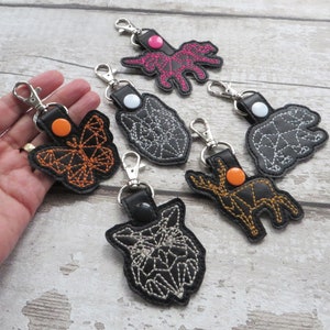 May include: A collection of black leather keychains with various geometric animal designs, including a butterfly, bear, and wolf. Each keychain features a metal clasp and colorful stitching, with orange, pink, and white accents.