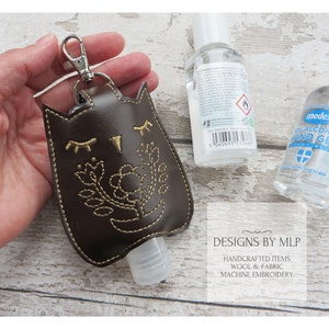 Könnte beinhalten: Brauner Leder-Handdesinfektionsmittel-Halter in Form einer Eule mit gestickten Blumen. Der Halter hat einen goldenen Reißverschluss und eine durchsichtige Plastikflasche mit Handdesinfektionsmittel. Der Text "DESIGNS BY MLP" ist auf dem Bild zu sehen.