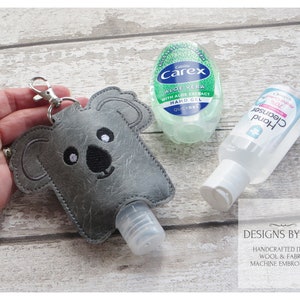 Può includere: Un porta-igienizzante per mani a forma di koala grigio con chiusura argentata, accanto a due flaconi di igienizzante per mani. Un flacone è verde con il testo "Carex Aloe Vera", l'altro è trasparente con il testo "Hand Cleanser".