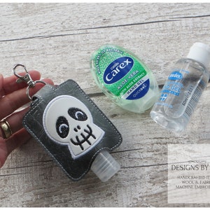Puede incluir: Un porta-gel hidroalcohólico gris en forma de calavera con un llavero plateado. El porta-gel es de tela y tiene un diseño de calavera blanca con ojos negros y boca. El porta-gel está unido a una pequeña botella de gel hidroalcohólico transparente. La imagen también muestra dos botellas de gel hidroalcohólico, una con una etiqueta verde y la otra con una etiqueta azul.