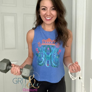 May include: A woman wearing a blue sleeveless shirt with a floral design and a crown. She is holding a grey dumbbell in one hand and a white beaded bracelet in the other. The shirt has the text "Grit & Grace Couture" on the bottom.