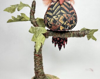 May include: A handmade owl ornament sitting on a decorative tree branch. The owl has a patterned body in black, orange, and brown, with pink feathery details. The tree has green leaves and a textured trunk, on a wooden base.