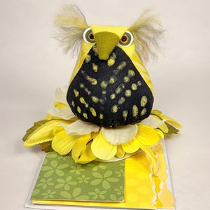 May include: A handmade yellow owl-shaped plush toy with a black and yellow patterned chest, large button eyes, and fluffy ear tufts. The owl sits atop a bed of yellow and white fabric flower petals, with a green floral patterned card below.