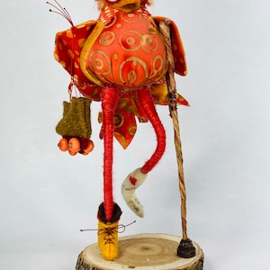 May include: A whimsical, handmade, orange and yellow bird doll with long, red legs and a brown walking stick. The bird is wearing a brown boot and has a small bag hanging from its leg. The bird is standing on a wooden log.