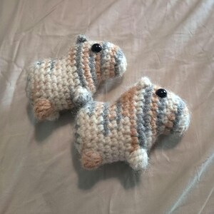 May include: Two handmade crochet capybaras. Each capybara is beige, gray, and peach with black button eyes. The capybaras are small, measuring approximately 3 inches in length, and have a soft, textured appearance.