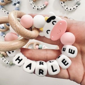 May include: A wooden teething ring with a cow-shaped silicone toy and a string of white silicone beads with black letters spelling out the name "HARLEE".