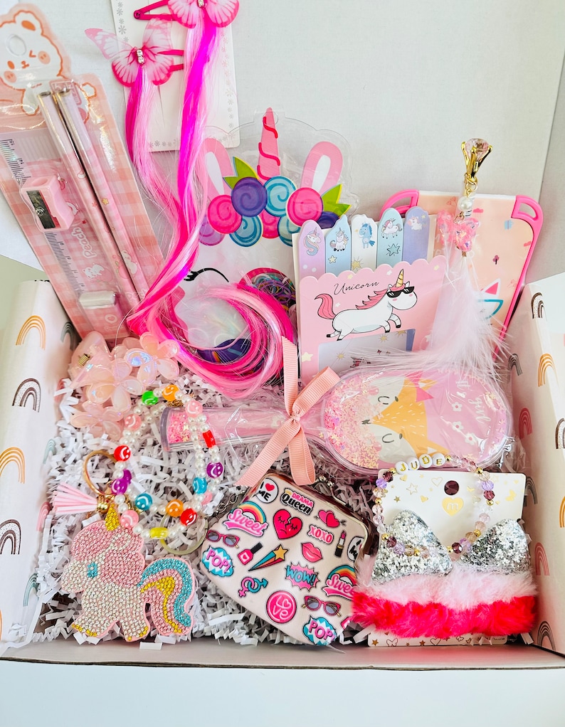 May include: A gift box containing unicorn-themed accessories. Items include a pink pencil set, hair clips with pink extensions, a unicorn keychain, a bracelet, a coin purse, and a sleep mask. The box is lined with white paper and decorated with rainbow and unicorn designs.