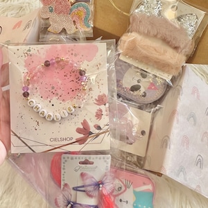 May include: A selection of children's accessories, including a bracelet spelling "CHARLIE", unicorn hair clips, and a glittery headband. The items are packaged in clear plastic and a brown paper bag with the "CIELSHOP" logo.