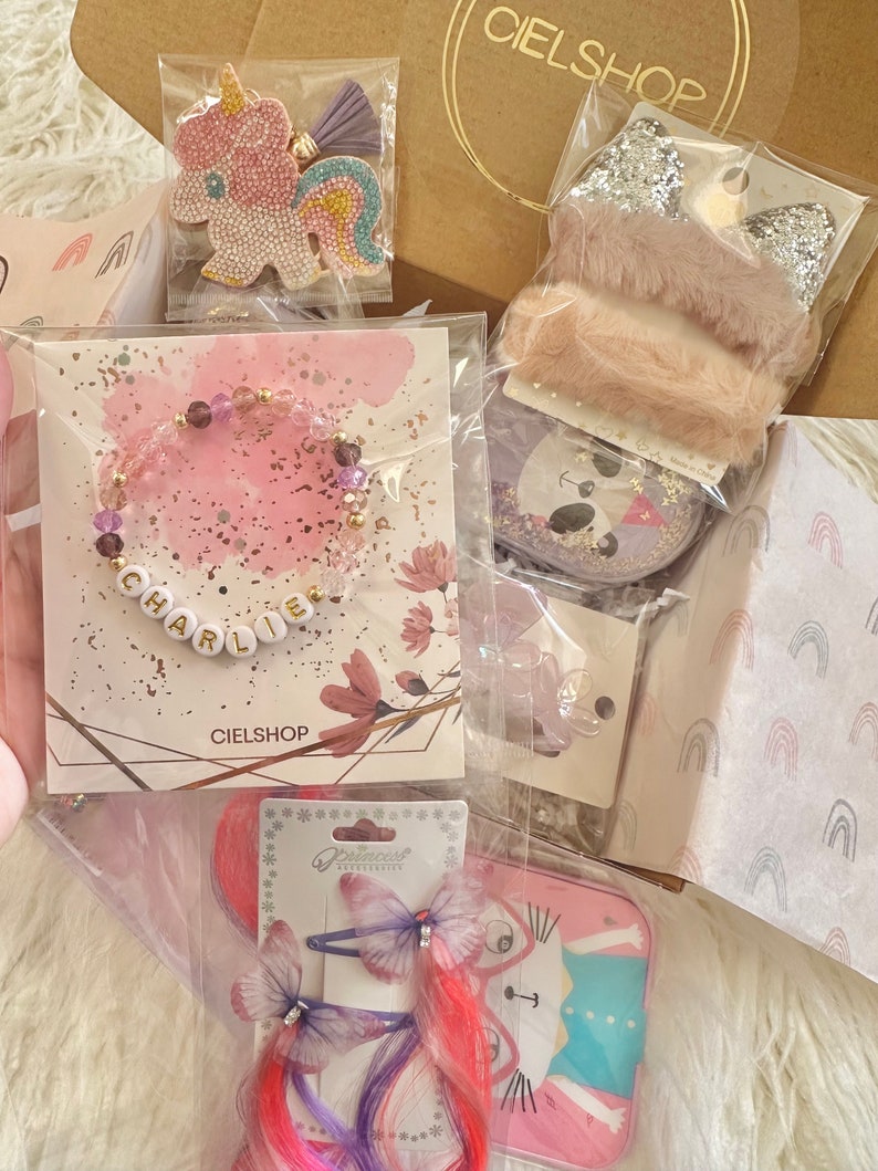 May include: A selection of children's accessories, including a bracelet spelling "CHARLIE", unicorn hair clips, and a glittery headband. The items are packaged in clear plastic and a brown paper bag with the "CIELSHOP" logo.