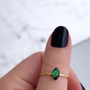 May include: A thin gold-toned ring featuring a small, marquise-shaped green stone set horizontally. The stone has a faceted surface with a deep emerald hue and a reflective quality. The band is smooth, slender, and metallic, encircling the finger.