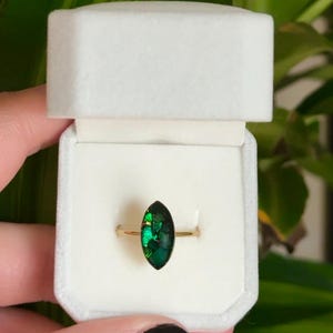 May include: A gold ring with a marquise-cut, iridescent green gemstone, presented in a white ring box. The gemstone reflects shades of green and gold. The ring is set against a blurred green background.