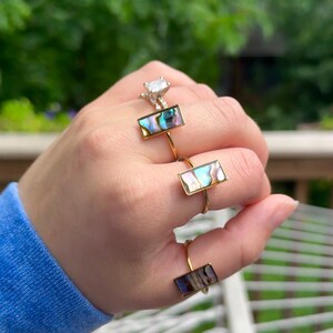 May include: Three gold-toned rings, each featuring a rectangular inlay of iridescent abalone shell. The rings have thin, simple bands and are worn on three separate fingers, showcasing the natural color variations of the shell in shades of blue, green, and purple.