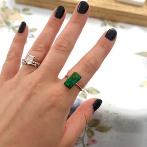 May include: A gold ring featuring a rectangular, iridescent green and blue stone set on a thin band, worn alongside a second ring with a clear rectangular stone and a delicate, diamond-encrusted band on a separate finger.